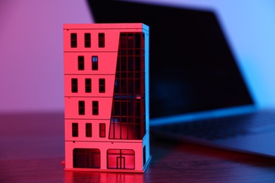 Photo of Real estate. Apartment building model and laptop on table in neon lights, closeup