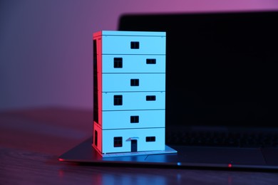 Photo of Real estate. Apartment building model and laptop on table in neon lights, closeup