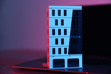 Photo of Real estate. Apartment building model and laptop on table in neon lights, closeup. Space for text