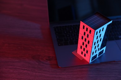 Photo of Real estate. Apartment building model and laptop on table in neon lights, closeup. Space for text