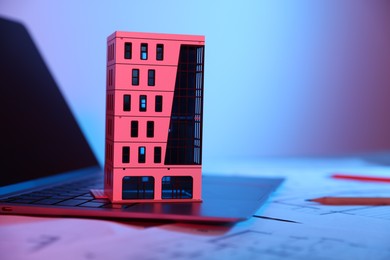 Photo of Real estate. Apartment building model, architectural drawings and laptop on table in neon lights, closeup. Space for text