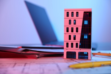 Photo of Real estate. Apartment building model, architectural drawings and laptop on table in neon lights, closeup