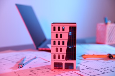 Photo of Real estate. Apartment building model, architectural drawings and laptop on table in neon lights, closeup