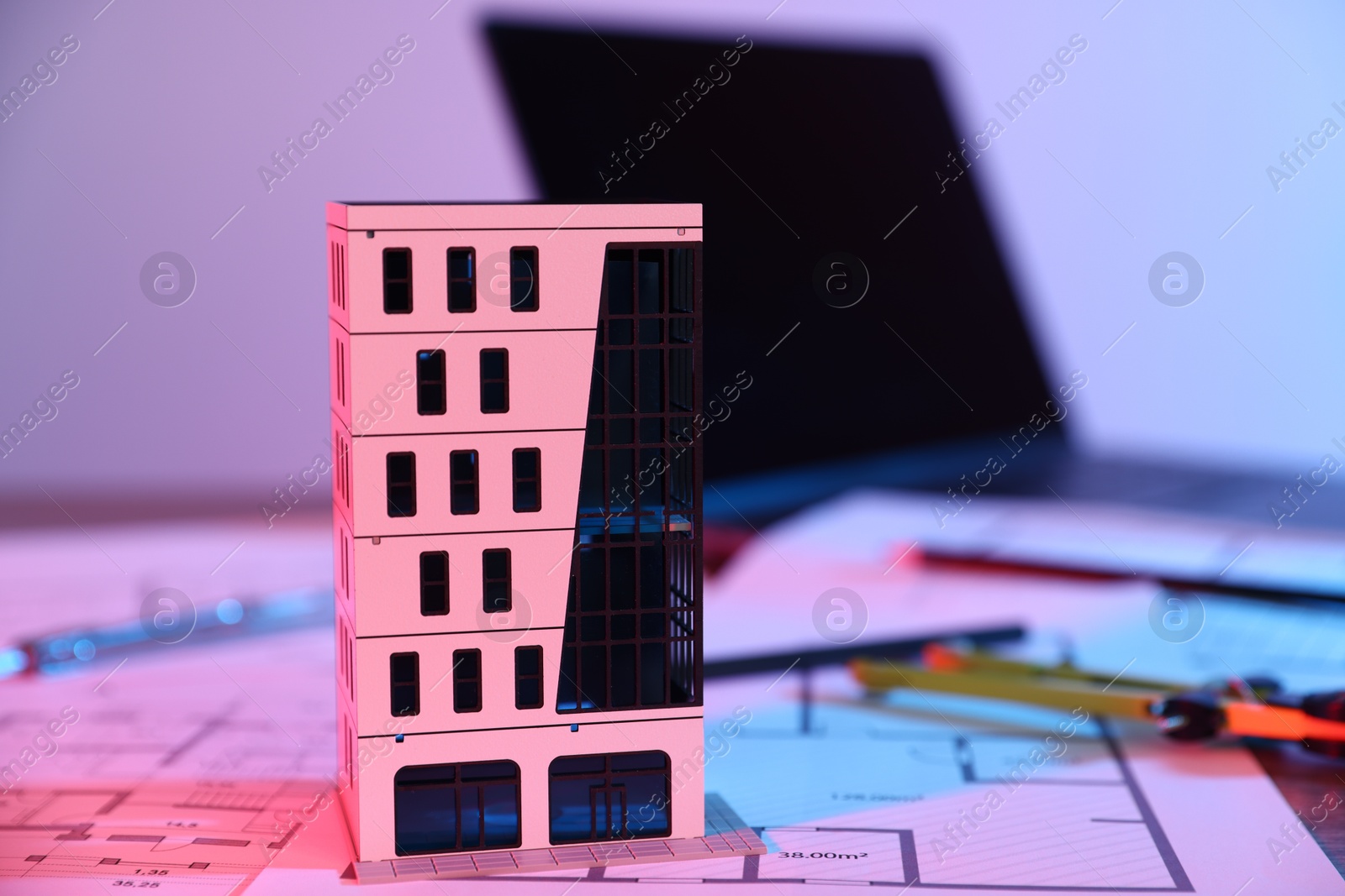 Photo of Real estate. Apartment building model, architectural drawings and laptop on table in neon lights, closeup
