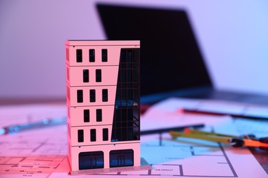 Photo of Real estate. Apartment building model, architectural drawings and laptop on table in neon lights, closeup