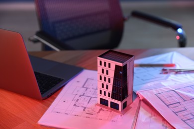 Photo of Real estate. Apartment building model, architectural drawings and laptop on table in neon lights, closeup
