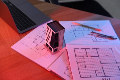 Photo of Real estate. Apartment building model, architectural drawings and laptop on table in neon lights, closeup