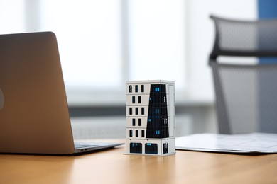 Photo of Real estate. Apartment building model, paperwork and laptop on table indoors, closeup