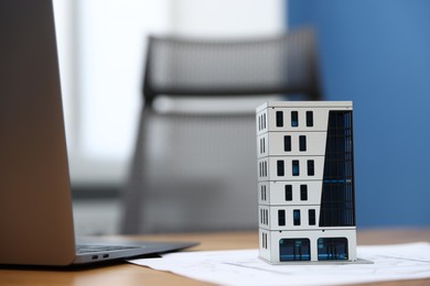 Photo of Real estate. Apartment building model, paperwork and laptop on table indoors, closeup