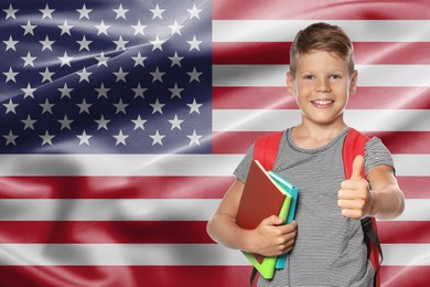 Image of Little boy with backpack and stationery showing thumbs-up against flag of USA. Language school