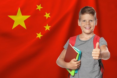 Image of Little boy with backpack and stationery showing thumbs-up against flag of China. Language school
