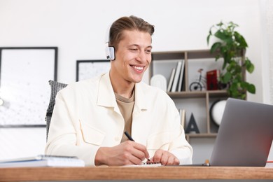 Photo of Man in headphones taking notes while passing online test via laptop at table indoors