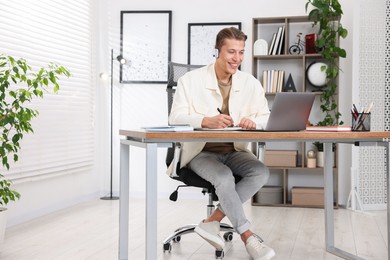 Photo of Man in headphones taking notes while passing online test via laptop at table indoors