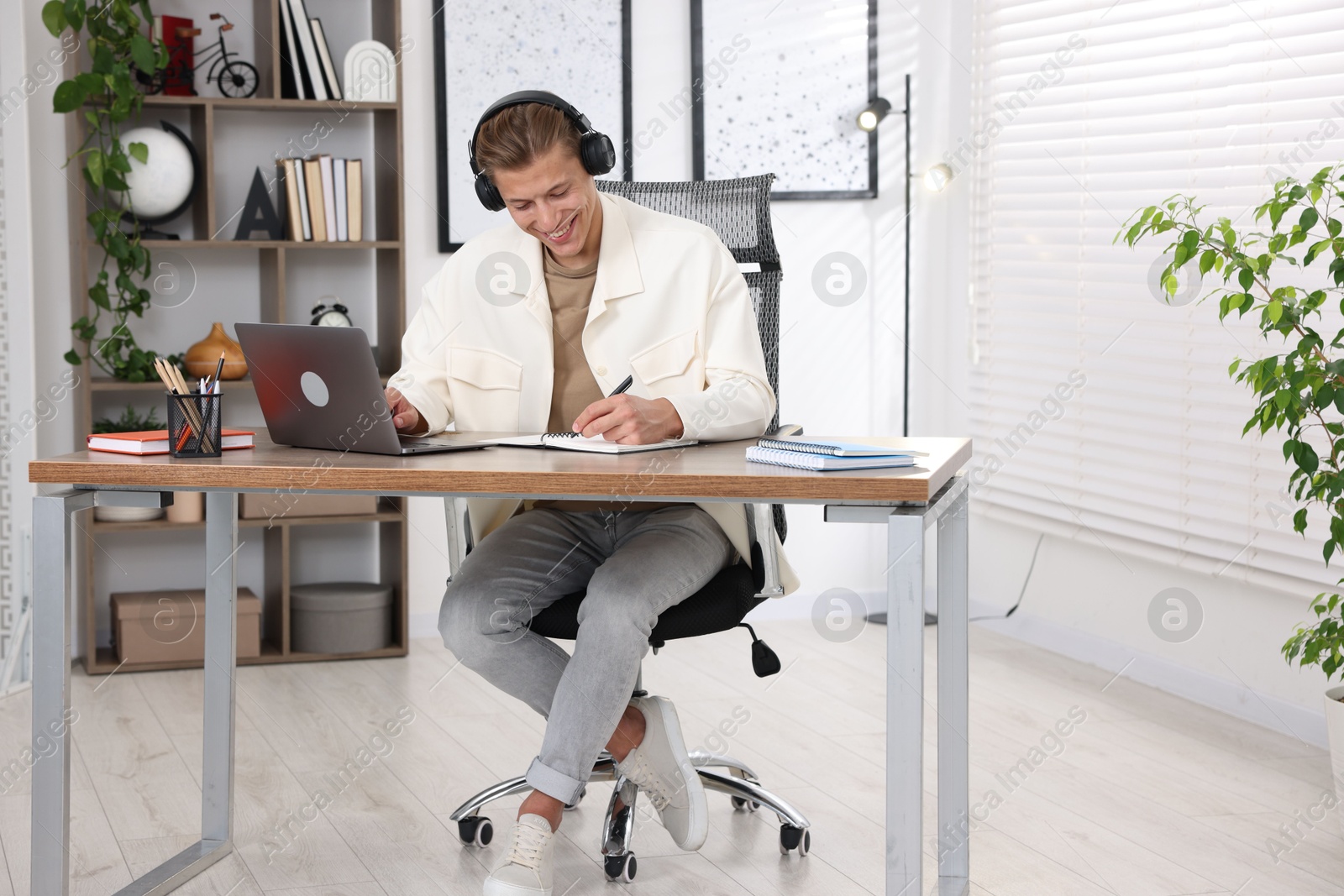 Photo of Man in headphones taking notes while passing online test via laptop at table indoors