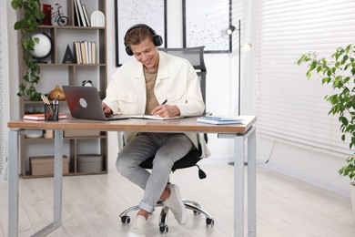 Photo of Man in headphones taking notes while passing online test via laptop at table indoors