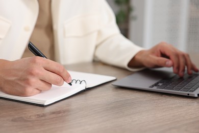 Photo of Man taking notes while passing online test on laptop at wooden table indoors, closeup