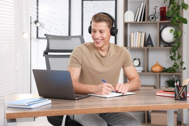 Photo of Man in headphones taking notes while passing online test via laptop at table indoors