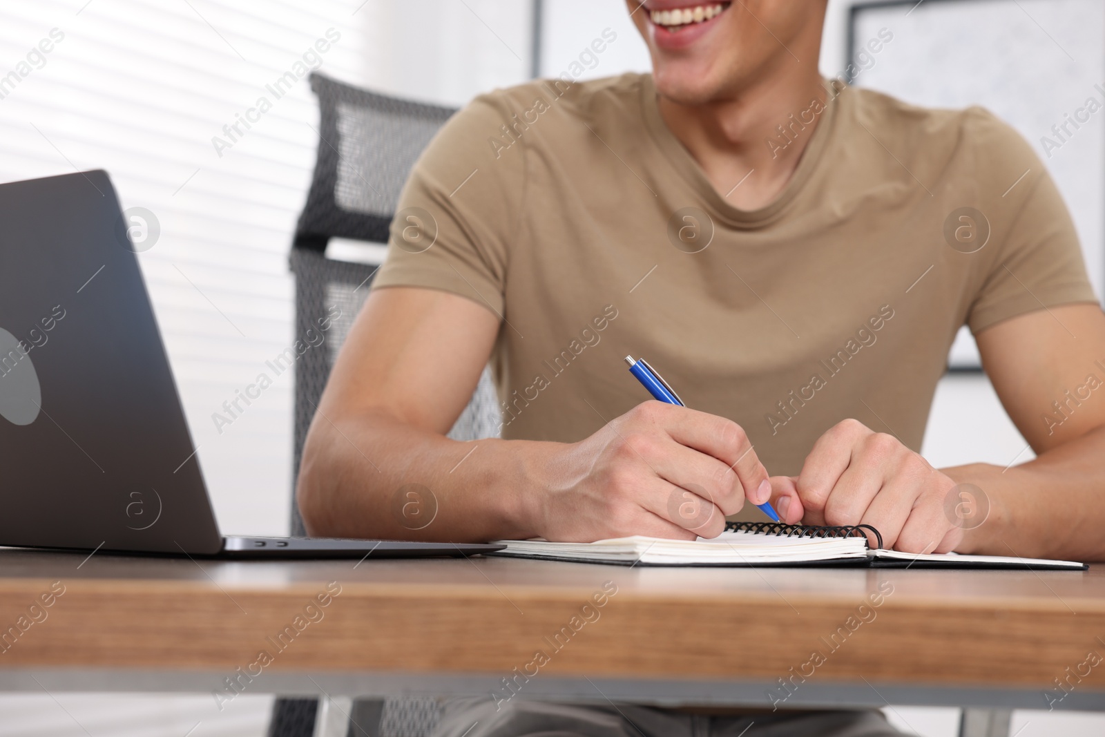 Photo of Man taking notes while passing online test via laptop at table indoors, closeup