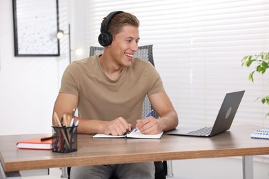 Photo of Man in headphones taking notes while passing online test via laptop at table indoors