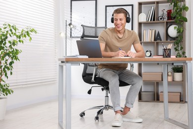 Photo of Man in headphones taking notes while passing online test via laptop at table indoors