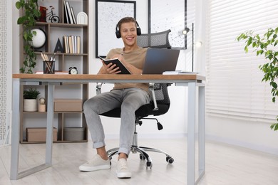 Photo of Man in headphones taking notes while passing online test via laptop at table indoors