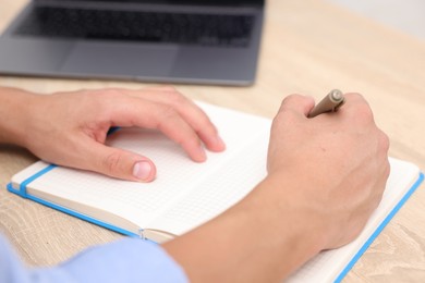 Photo of Man taking notes while passing online test via laptop at wooden table indoors, closeup