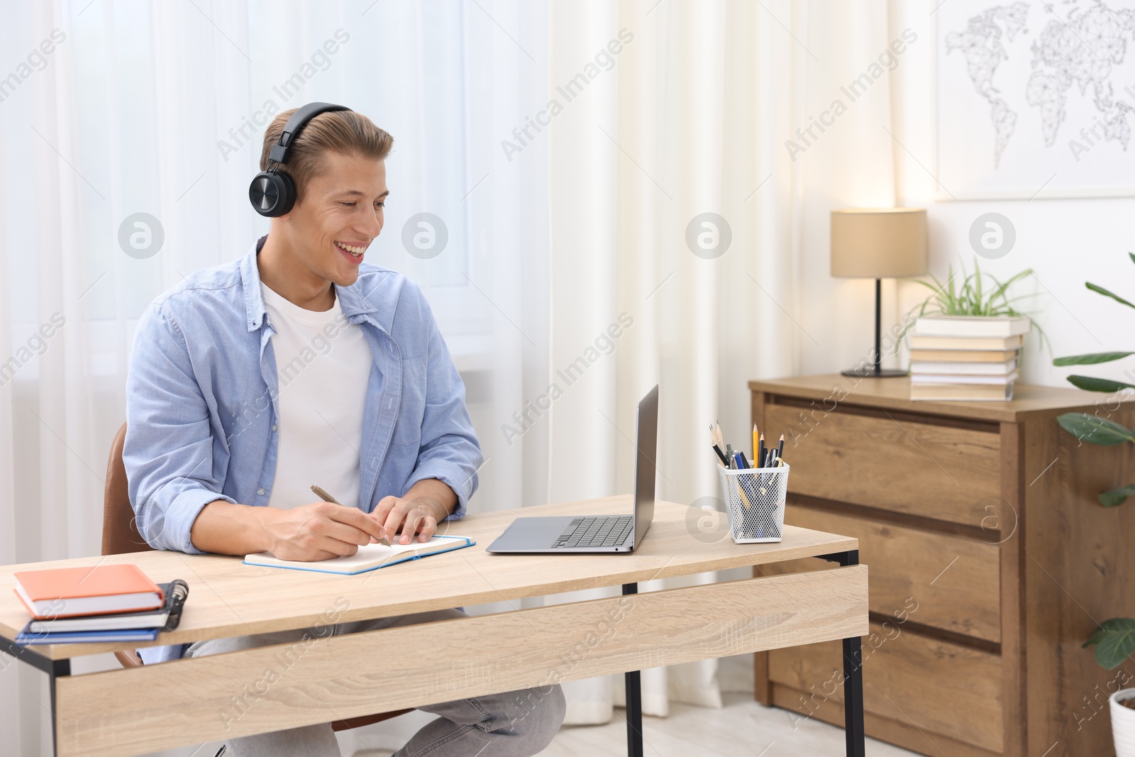 Photo of Man in headphones taking notes while passing online test via laptop at table indoors