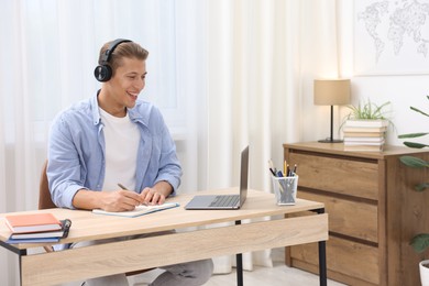 Photo of Man in headphones taking notes while passing online test via laptop at table indoors