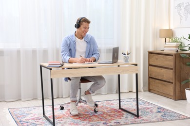 Photo of Man in headphones taking notes while passing online test via laptop at table indoors