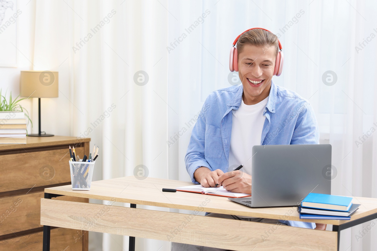 Photo of Man in headphones taking notes while passing online test via laptop at table indoors