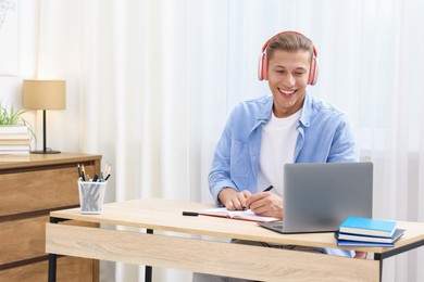 Photo of Man in headphones taking notes while passing online test via laptop at table indoors