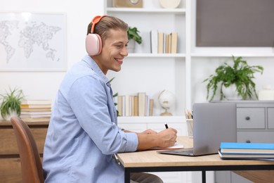 Photo of Man in headphones taking notes while passing online test via laptop at table indoors
