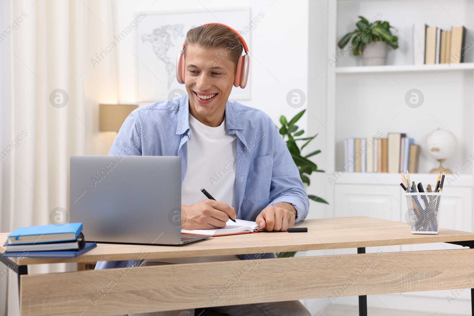 Photo of Man in headphones taking notes while passing online test via laptop at table indoors