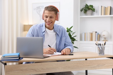 Photo of Man in headphones taking notes while passing online test via laptop at table indoors