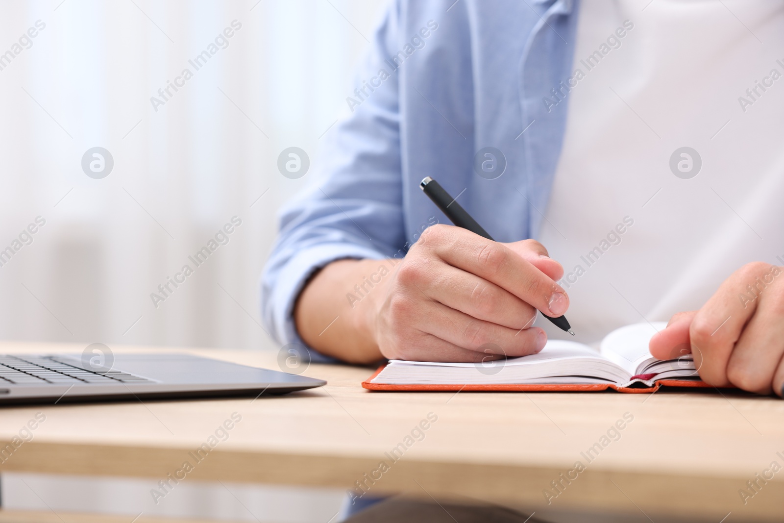 Photo of Man taking notes while passing online test via laptop at wooden table indoors, closeup