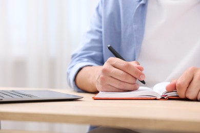 Photo of Man taking notes while passing online test via laptop at wooden table indoors, closeup