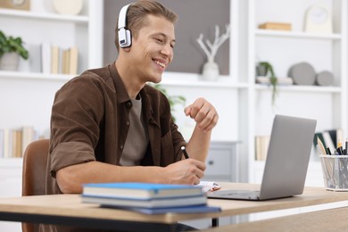 Photo of Man in headphones taking notes while passing online test via laptop at table indoors