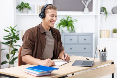 Photo of Man in headphones taking notes while passing online test via laptop at table indoors