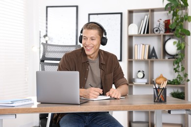 Photo of Man in headphones taking notes while passing online test via laptop at table indoors