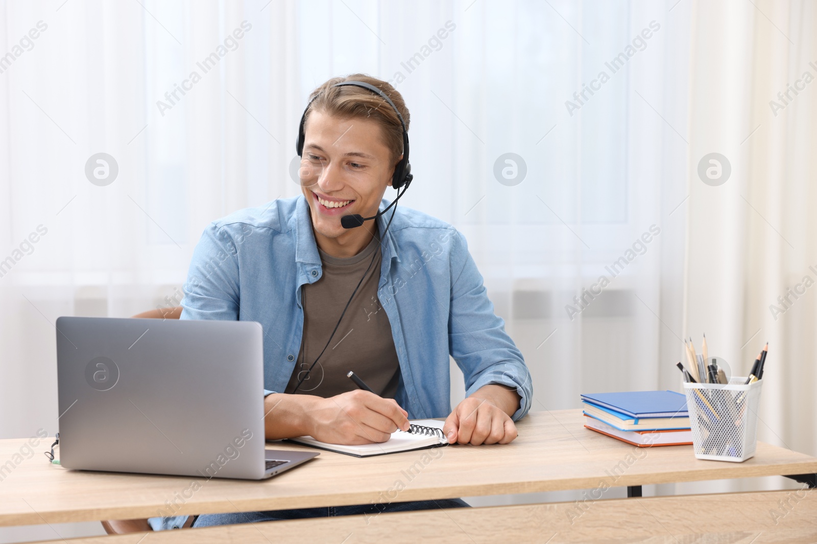 Photo of Man in headset taking notes while passing online test via laptop at table indoors