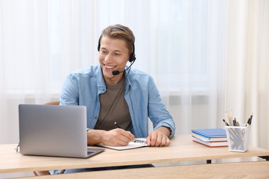 Photo of Man in headset taking notes while passing online test via laptop at table indoors