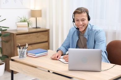 Photo of Man in headset taking notes while passing online test via laptop at table indoors