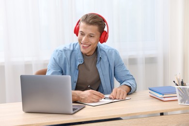 Photo of Man in headphones taking notes while passing online test via laptop at table indoors