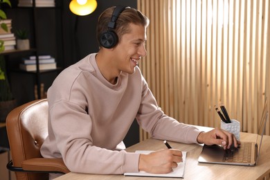 Photo of Man in headphones taking notes while passing online test via laptop at table indoors