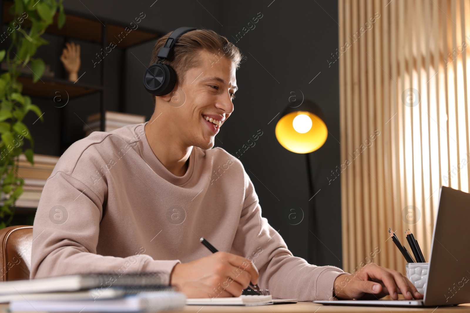 Photo of Man in headphones taking notes while passing online test via laptop at table indoors
