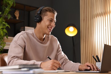 Photo of Man in headphones taking notes while passing online test via laptop at table indoors