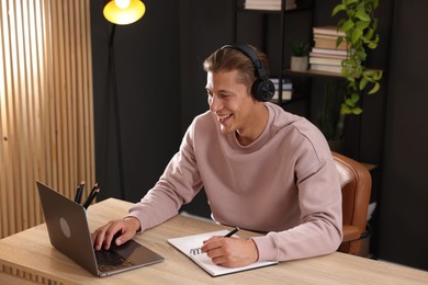Photo of Man in headphones taking notes while passing online test via laptop at table indoors
