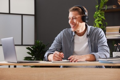 Photo of Man in headphones taking notes while passing online test via laptop at table indoors