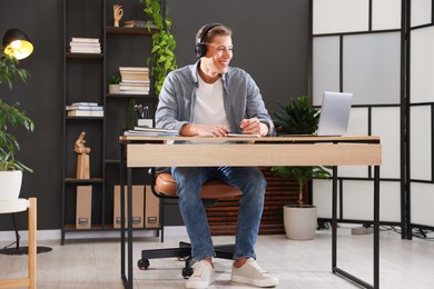Photo of Man in headphones taking notes while passing online test via laptop at table indoors