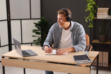 Photo of Man in headphones taking notes while passing online test via laptop at table indoors